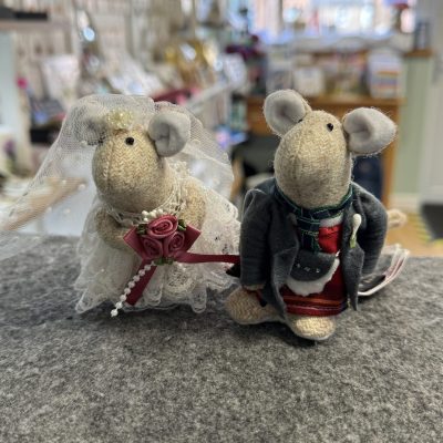 Two charming handmade wedding mice figurines are displayed on a gray surface in a bright, cozy gift shop. One felt mouse features a delicate bridal veil and holds a red bouquet, while the other wears an elegant groom’s suit with a classic bow tie—perfect as unique wedding cake toppers or thoughtful handcrafted wedding decor.