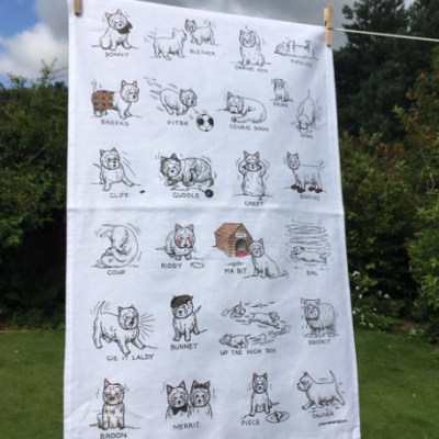 A charming West Highland Terrier tea towel is prominently displayed against a backdrop of lush greenery and a bright blue sky. This delightful Westie dog tea towel features illustrated West Highland Terriers alongside popular Scottish dialect words such as cannae and braw. Each term is creatively paired with enchanting depictions of dog activities, making this tea towel a must-have for any dog lover or Scottish culture enthusiast.