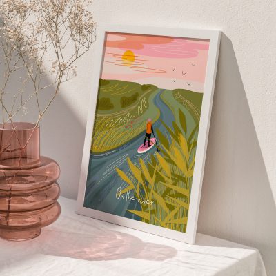 An A4 art print from the On the River collection complements a pink glass vase filled with dried flowers. This stunning art piece beautifully captures a person paddleboarding on a river at sunset, surrounded by picturesque hills and birds in flight. The artworks title is elegantly inscribed across the scene, making it an ideal addition for any art lover or home decor enthusiast seeking unique wall art.