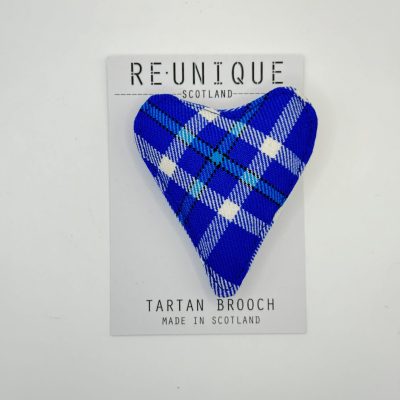 Introducing a Scottish tartan brooch designed in the shape of a flower, featuring an eye-catching blue, white, and black pattern accented with light purple hues. This exquisite brooch is presented on a card elegantly branded with REUNIQUE SCOTLAND and proudly stating TARTAN BROOCH MADE IN SCOTLAND.