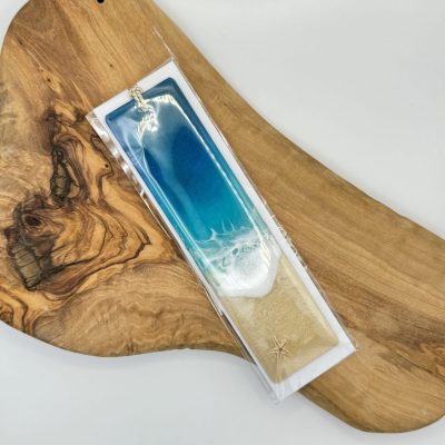The Resin Waves Bookmark elegantly rests on a wooden cutting board, showcasing a stunning ocean-like gradient transitioning from blue to white. This eye-catching bookmark features a warm tan base and is beautifully accented with a charming starfish decoration, making it the perfect coastal-themed accessory.