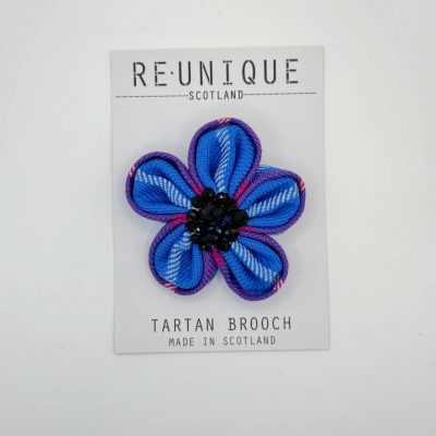 Displayed on a white card with the inscription REUNIQUE SCOTLAND Tartan Brooch Made in Scotland, this exquisite brooch features a light purple, flower-shaped design with five petals. The center of this Scottish tartan brooch is elegantly accented with black bead details, making it a perfect accessory for any ensemble.