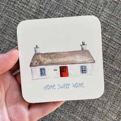 A Bothy Coaster showcasing a charming cottage with a thatched roof, twin chimneys, and a vibrant red door is adorned with the phrase Home Sweet Home. Perfect for enhancing your home décor, this coaster design sits elegantly on a textured gray surface.