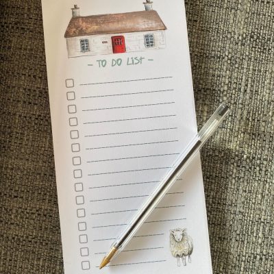 The Bothy To-Do List Pad is perfect for organizing your tasks with its charming design. Featuring blank checkboxes, an enchanting cottage illustration at the top, and an adorable sheep graphic at the bottom, this pad is both functional and visually appealing. Complemented by a stylish clear pen with a silver tip placed diagonally across it, this set creates a captivating contrast against a textured gray fabric background. Make productivity enjoyable with this beautiful to-do list pad and sleek pen combo!.