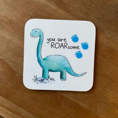 The Dino Coaster, featuring a light blue design, showcases a cartoon dinosaur alongside the playful phrase you are ROARsome on a wooden background. Enhance your décor with this charming coaster, adorned with three small blue footprints above the dino, perfect for dinosaur lovers and home accessory enthusiasts seeking whimsical and unique designs.