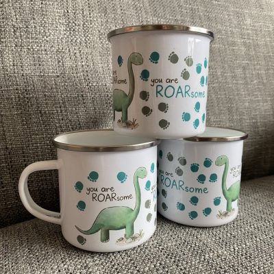 Three Stacked Dinosaur Enamel Mugs - You Are ROARsome | Green Dinosaur Illustrations with You Are ROARsome Text on Rustic Gray Textured Surface and Playful Blue and Green Dino Footprints.