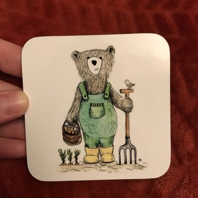 The Garden Bear Coaster showcases a charming bear in green overalls, holding a basket of carrots and a garden fork, beautifully decorated with a small bird. This delightful coaster sits on a red-brown textured background adorned with carrot plants at its feet, making it perfect for enhancing garden-themed décor.
