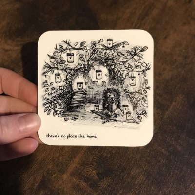 A hand showcases the Hedgehog Home Coaster, highlighting a detailed tree adorned with lanterns and a hedgehog nestled among lush foliage. The coaster features the heartfelt phrase Theres No Place Like Home, all beautifully set against a rich, dark wooden background. Perfect for nature-themed decor or as a charming home accessory, this hedgehog coaster adds warmth and personality to your living space.