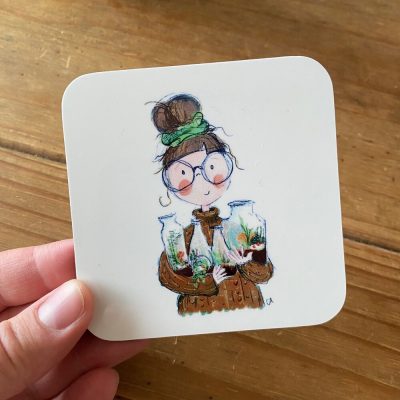 A hand showcases a Terrarium Lady Coaster, adorned with an illustration of a woman sporting glasses and a bun, dressed in a brown sweater, and gently holding three terrariums filled with miniature plants.