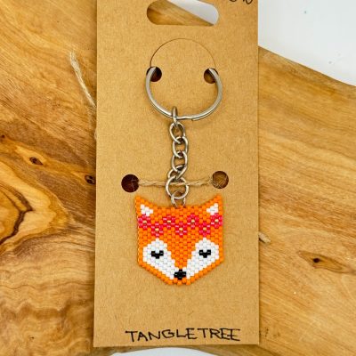 Introducing a handcrafted beaded keychain showcasing an adorable orange fox head with white facial details and striking black eyes. This charming fox keychain is securely attached to a brown cardboard backing, displaying the text £10 and TANGLE TREE. The natural wooden surface backdrop adds a rustic touch to this unique beaded accessory. Perfect for animal lovers and those seeking distinctive handmade gifts!.
