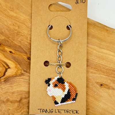 Discover our handcrafted beaded guinea pig keychain, elegantly displayed on a rustic wooden surface. This charming accessory, presented on a Tang Le Tree card, can be yours for just £10. Perfect for animal lovers and collectors alike!.