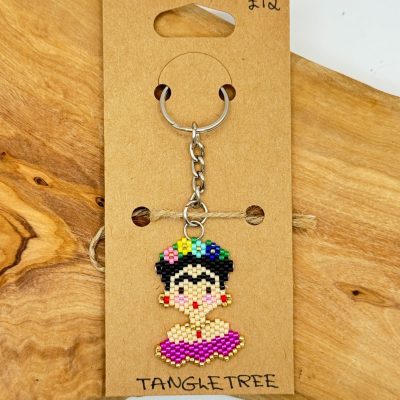 A vibrant handmade keychain featuring a beaded figurine adorned in colorful clothing and a floral headband is attached to a brown card labeled TANGLETREE. This eye-catching accessory, priced at £12, sits on a rustic wooden surface.