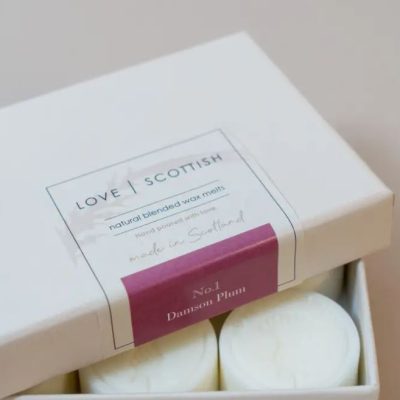 A creatively designed box labeled LOVE SCOTTISH offers premium natural blended wax melts in the delightful No.1 Damson Plum scent. The packaging proudly showcases messages such as hand poured with love and made in Scotland. Inside, you can partially see three elegant white wax melts ready to infuse your space with their captivating aroma.