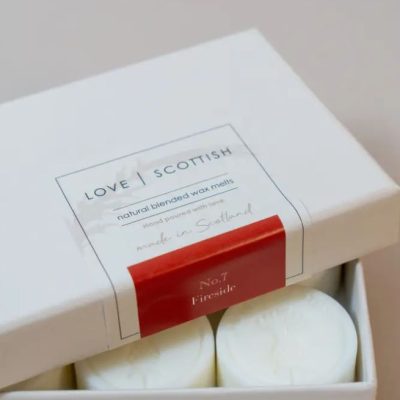 A close-up image showcases a box labeled Love Scottish: Natural Blended Wax Melts. This white box with a striking red label highlights the popular scent No. 7 Fireside. Two white wax melts can be seen partially inside, illustrating their inviting fragrance and premium quality.