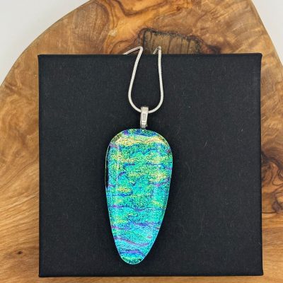 Discover the enchanting allure of an iridescent teardrop pendant in mesmerizing shades of blue, green, and purple. Suspended on a premium sterling silver chain, this exquisite jewelry piece gracefully rests against a striking black square background atop a natural wooden surface.