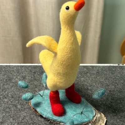 A charming felt duck figurine with vibrant red boots stands gracefully on a faux pond crafted from high-quality felt and wood. Surrounding this delightful scene are small, meticulously designed felt plants. This picturesque setup is perfectly complemented by a neutral-colored curtain backdrop, making it an ideal centerpiece for home decor enthusiasts and craft lovers alike.