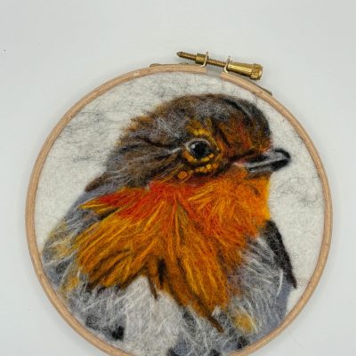 Discover a stunning felted wool artwork of a robin showcased in an embroidery hoop. This masterpiece features the robin with bright orange and rich brown feathers set against a soft cream background, perfect for bird lovers and home décor enthusiasts seeking unique artistry.