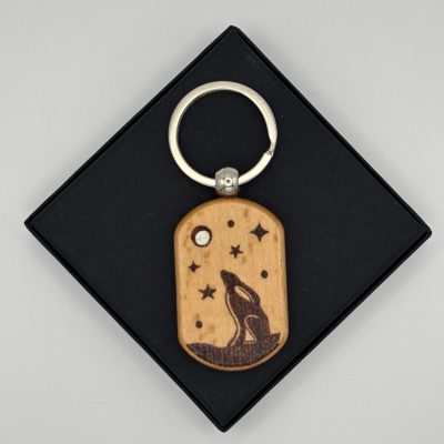 This eye-catching keychain boasts a unique wooden design paired with a round metal keyring. It highlights an intricately engraved howling wolf, encircled by stars and a moon, offering the sophistication of a Sterling Silver Hammered Bangle. The design is beautifully displayed against a black square backdrop, making it both stylish and functional for everyday use.