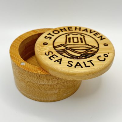 Discover the elegance of a wooden salt container, expertly crafted with a smooth, natural finish. This exquisite storage piece features an engraving of Stonehaven Sea Salt Co. alongside a detailed image of a lighthouse. With its lid slightly open, this container not only offers functionality but also adds a touch of coastal charm to your kitchen décor. Perfect for preserving the flavor and quality of your sea salt collection.