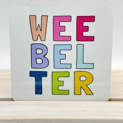 Vibrant letters spell WEE BELTER against a square white backdrop, set on a light wood surface.