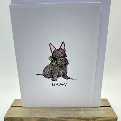 Greeting card showcasing an illustration of a black dog with pointy ears and a stylish plaid collar. The text BRAW is featured beneath the dog. The card is elegantly presented on a wooden stand against a simple background.