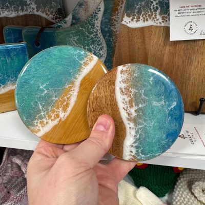 A hand holds two ocean wave-themed round coasters, expertly designed to mimic the serene meeting of waves and the shoreline. These beach-inspired coasters showcase a stunning blend of blue, white, and natural wood colors. In the background, you can see more coastal decor items that enhance any seaside home aesthetic.