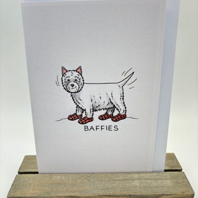 Illustration of a dog wearing red plaid slippers on a piece of paper titled BAFFIES, propped up on a wooden stand against a minimalist backdrop.
