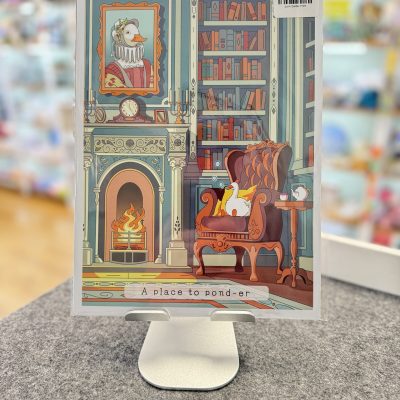 Illustration of a cozy library featuring a duck in an armchair by the fireplace, surrounded by bookshelves and art depicting another duck. The display stand highlights the charming text, A Place to Pond-er.