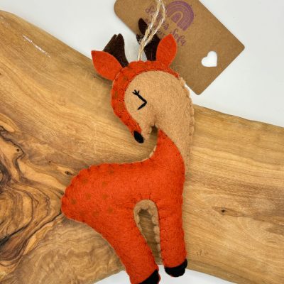 Discover our charming handmade felt deer ornament, featuring a vibrant red-orange body, tan face, and black hooves. This adorable felt deer decoration is perfect for adding a touch of whimsy to any space. Its closed eyes lend a peaceful vibe, and it comes with an attached string for easy hanging. This unique ornament also includes a brown tag adorned with a heart logo, enhancing its handcrafted appeal. Perfect for holiday decor or as year-round woodland-themed accents!.