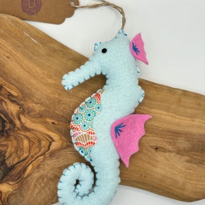 Displayed on a wooden board is a beautiful handmade felt seahorse featuring vibrant blue and patterned fabric. This unique piece is accented with pink felt fins and detailed blue stitching. A rustic brown tag with personalized writing is attached to the seahorse, making it an ideal decor item for ocean-themed rooms or as a thoughtful handmade gift.