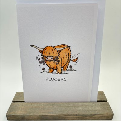 A charming greeting card displayed on a wooden stand showcases an illustration of a Highland cow nestled in grassy meadows adorned with vibrant flowers. The bold caption FLOOERS stands prominently beneath the image, adding a touch of rustic charm.