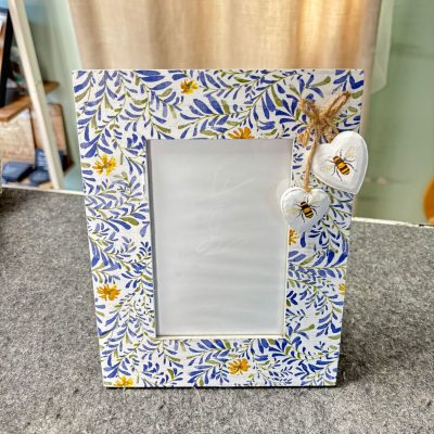 Introducing a stunning decorative photo frame featuring a beautiful floral pattern in blue and yellow, perfect for enhancing your home decor. This charming frame sits elegantly on a grey surface, adorned with two small heart ornaments showcasing intricate bee illustrations, tied with rustic twine in the top right corner. The backdrop of this scene includes stylish curtains and thoughtfully curated shelves, adding to the overall aesthetic appeal.
