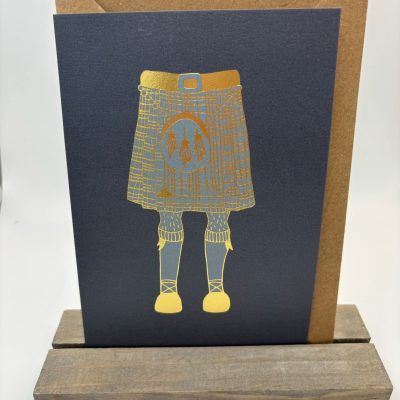 Discover our premium greeting card featuring a stunning gold-embossed illustration of a kilt and sporran set against a rich dark background. This elegant card is beautifully displayed on a wooden stand, with an attractive brown envelope subtly peeking out behind it. Perfect for any occasion, this eye-catching design is sure to leave a lasting impression.