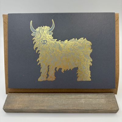 This elegant greeting card showcases a luxurious gold-embossed illustration of a fluffy Highland cow set against a rich dark background. Perfectly displayed on a minimalist wooden stand, this card is sure to catch the eye and bring joy to any recipient.