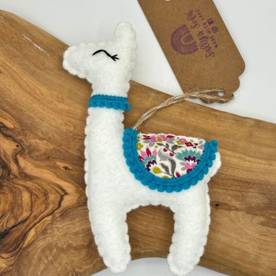 Discover our handcrafted plush llama, featuring a vibrant design perfect for any decor. This unique white plush llama showcases a colorful saddle and eye-catching blue trim. Designed for convenience, it includes a thread loop for easy hanging against a wooden board background. A stylish brown tag adds the final touch to this delightful llama decoration. Perfect as a gift or charming detail to brighten your space!.