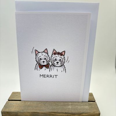 A charming greeting card is showcased on a wooden stand, featuring an adorable illustration of two fluffy dogs. The word MERRIT is elegantly displayed beneath the cute canine duo. The design is set against a plain background, perfect for those seeking unique dog-themed cards or personalized greeting cards with charming pet illustrations.