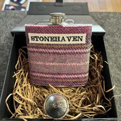 A stylish metal flask wrapped in a vibrant purple and pink patterned fabric labeled STONEHAVEN is elegantly displayed in a black gift box filled with decorative straw. The open gift box showcases the flask alongside its round, shiny cap, all set against a rustic wood floor backdrop. This eye-catching presentation makes for a perfect decorative liquor accessory or thoughtful gift idea.