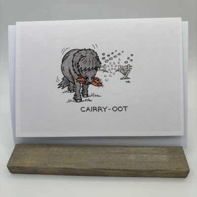 A humorous greeting card features a cartoon sheepdog walking upright with food in its mouth. The caption below playfully spells CAIRRY-OOT. This funny greeting card is attractively displayed on a wooden stand.