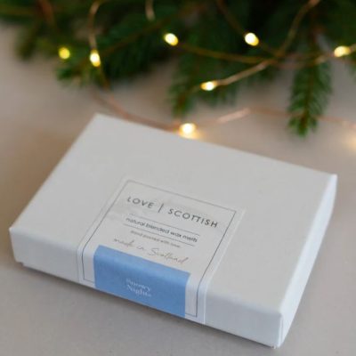 A Love Scottish white box featuring natural wax melts sits elegantly on a table, perfect for those seeking eco-friendly scents. The box showcases a stylish blue strip labeled Snowy Night, ideal for creating a cozy winter ambiance. Soft, warm fairy lights and vibrant green foliage provide a blurred background, enhancing the inviting atmosphere.