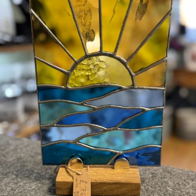 Discover a stunning stained glass art piece on a wooden stand, showcasing a breathtaking sunset over serene blue waves. This exquisite artwork features vibrant colors with yellow and orange hues for the radiant sun and sky, seamlessly transitioning into varied shades of blue representing calm waters. Perfect for collectors or as an eye-catching home decor accent, this unique stained glass creation includes an attached price tag for easy purchasing.