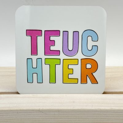 Vibrant, colorful letters spell TEUCHTER on a white square card, elegantly displayed on a light wooden stand.