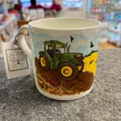 A ceramic mug featuring a vibrant illustration of a green tractor on a picturesque farm. The tractor drives through lush fields with birds soaring in the sky and chickens clucking nearby. To the right, bright yellow crops add a splash of color. Perfectly displayed on a gray surface, this charming farm-themed mug is an ideal addition to any store setting.