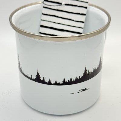 White enamel camping mug featuring a black silhouette forest and canoe design on a serene lake. This metal rimmed camper’s mug also holds a rolled-up accessory with stylish black and white stripes, perfect for outdoor enthusiasts.