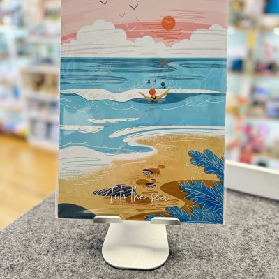 Displayed on a stand, this vibrant notebook cover showcases an eye-catching beach scene illustration. With rolling waves, a swimmer gliding through the water, and a stunning setting sun, it captures the essence of coastal beauty. Above, birds soar gracefully in the sky while the inspirational phrase Into the Sea elegantly adorns the bottom. Perfect for those seeking ocean-inspired stationery!.