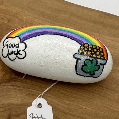 A charming painted rock with a white background showcases a vibrant rainbow, a pot of gold adorned with a green clover, and the term Good Luck beautifully scripted in a cloud. This unique decorative rock, tagged as #6, is artfully displayed on a wooden surface. Perfect for adding color and luck to your home decor or garden display.