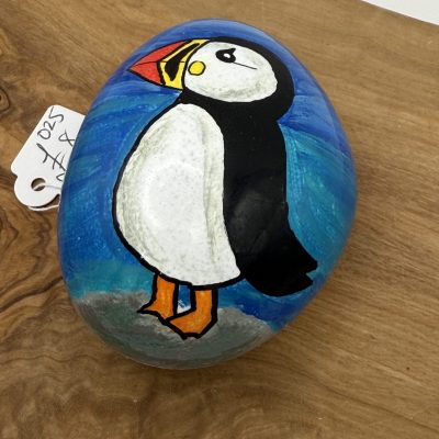 A beautifully hand-painted puffin stone featuring striking black and white feathers, a vibrant red and yellow beak, and distinctive orange feet on a serene blue background. This unique puffin art piece is displayed on a natural wooden surface, with a visible price tag indicating its value as a perfect gift or decorative item.