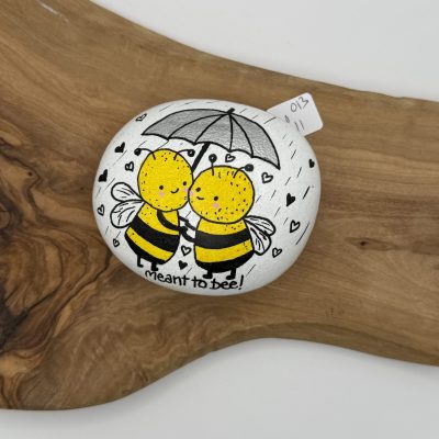 A hand-painted rock showcases two cheerful bees holding an umbrella, adorned with small heart accents. The inscription meant to bee! adds charm to this delightful creation. This decorative rock art rests on a rustic wooden surface, making it a perfect addition to your nature-inspired decor.