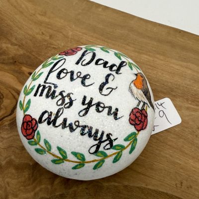 A hand-painted stone resting on a wooden surface displays the heartfelt message Dad love & miss you always, beautifully adorned with red roses and a charming small bird illustration. A small price tag is affixed to the right side of this memorial stone keepsake.