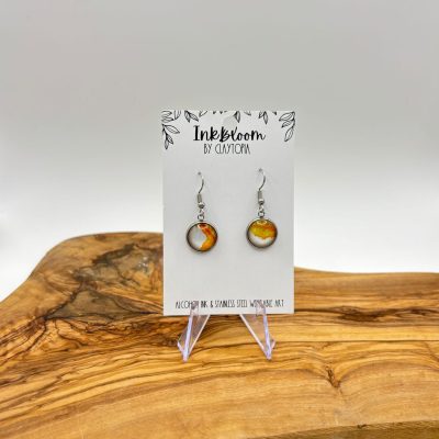 Discover the elegance of our Amber and White Inkbloom Earrings, featuring exquisite circular swirls of amber and yellow. These stunning earrings gracefully dangle from a white card labeled Inkbloom by Claytona, beautifully presented on a natural wood grain surface. Perfect for enhancing your fashion style with a touch of sophistication.