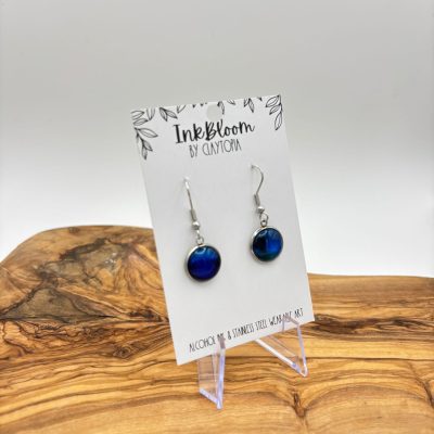 The Blue Inkbloom Earrings featuring silver hooks are elegantly showcased on a white InkBloom by ClayTotal card. These stunning earrings, resting in a clear holder on a wooden surface, create a beautiful contrast against the smooth, natural wood texture. Perfect for fashion enthusiasts seeking unique handmade jewelry.